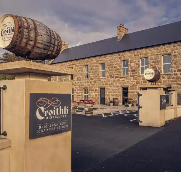Stone building with "Croithlà Distillery" signs in English and Irish, featuring a large barrel above the entrance gates. Enjoy outdoor seating beneath a cloudy sky and discover Crolly Distillery tours Donegal offers to visitors.