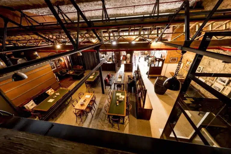 A spacious, rustic restaurant with wooden tables and chairs, warm lighting, and industrial-style decor—perfect for relaxing after Crolly Distillery tours in Donegal. A few people are inside, and framed pictures hang on the walls.