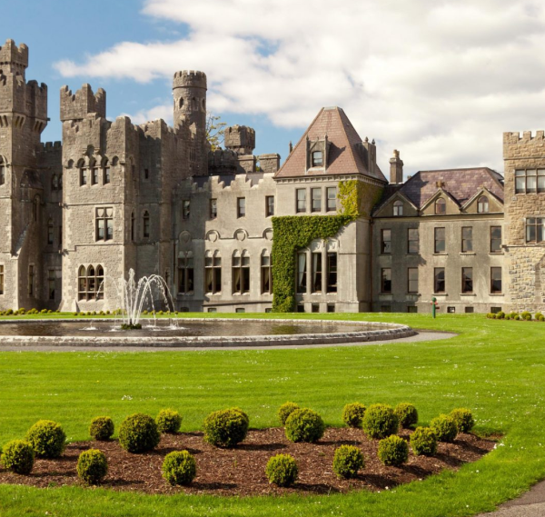 A large stone castle with turrets and towers stands behind a round fountain in a manicured lawn with trimmed shrubs. Sunlight brightens the historic building, a highlight of Ancient East tours Ireland under a partly cloudy sky.