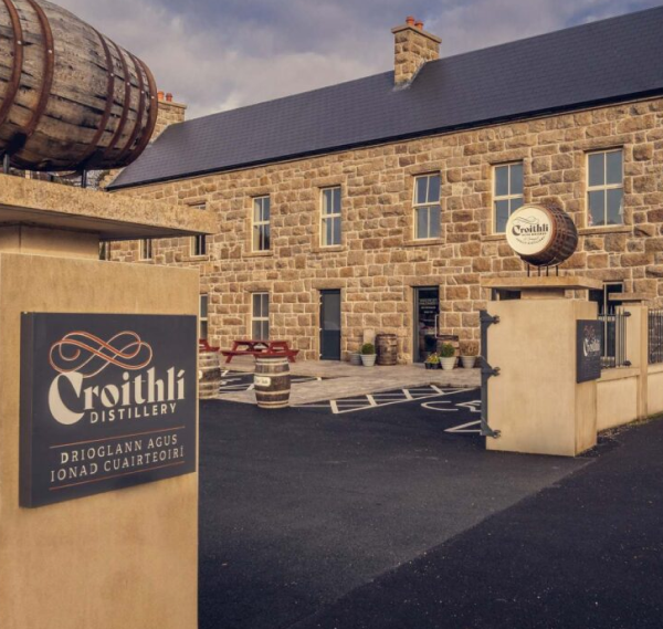 A stone building with the sign "Croithlí Distillery" in both Irish and English, featuring a large barrel above the gate, outdoor seating, accessible parking spaces, and a popular stop on Ancient East tours Ireland.