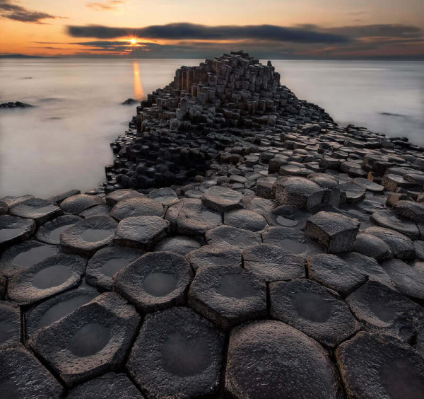 Geometric stone columns of the Giant’s Causeway lead into the calm sea at sunset, with golden light illuminating the clouds and water—a must-see highlight on Ancient East tours Ireland.