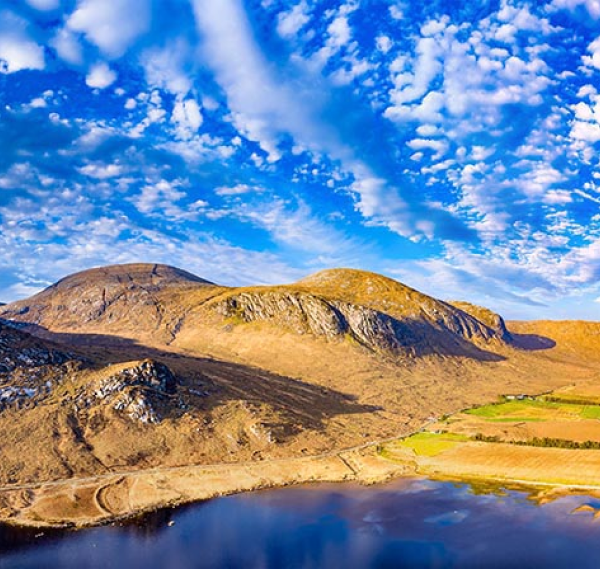 A bright landscape with sunlit hills, rocky slopes, green fields, and a winding road evokes the beauty often seen on Ancient East tours Ireland, with a still lake reflecting a vivid blue sky scattered with clouds.