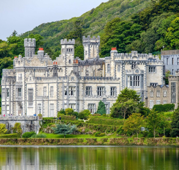An ancient stone castle with towers and battlements stands by a calm lake, surrounded by lush green hills—a perfect sight on Ancient East tours Ireland.