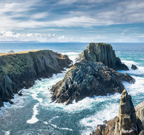 Rocky cliffs and jagged sea stacks rise from blue ocean waves under a partly cloudy sky, with green grass atop the cliffs—just one breathtaking scene to discover on Ancient East tours Ireland offers, with distant land visible on the horizon.