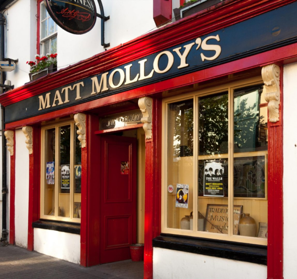 The exterior of Matt Molloy's pub with a red-trimmed façade, large windows displaying posters, and a sign featuring the pub's name above the entrance is a highlight for visitors enjoying Ancient East tours Ireland.