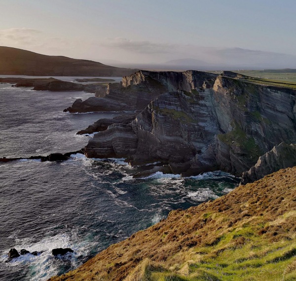 Steep rocky cliffs covered in patches of grass rise above the sea, with waves crashing below—an iconic scene featured on Ancient East tours Ireland, where the coastline curves into the distance under a clear, softly lit sky.