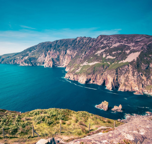 Steep sea cliffs covered in greenery rise above deep blue waters under a clear sky, with rugged rock formations stretching into the sea and lush grass in the foreground—an iconic view on Ancient East tours Ireland.