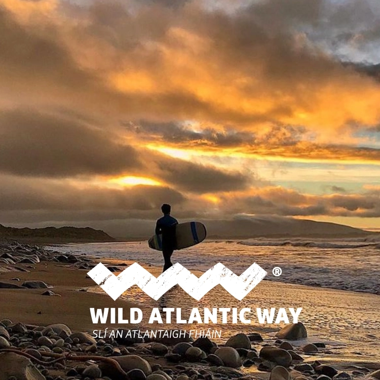 A surfer holding a surfboard walks along a rocky beach at sunset, with dramatic clouds and golden light in the sky. Text reads: "Wild Atlantic Way Slí an Atlantaigh Fhiáin – discover Donegal with unforgettable Wild Atlantic Way tours.