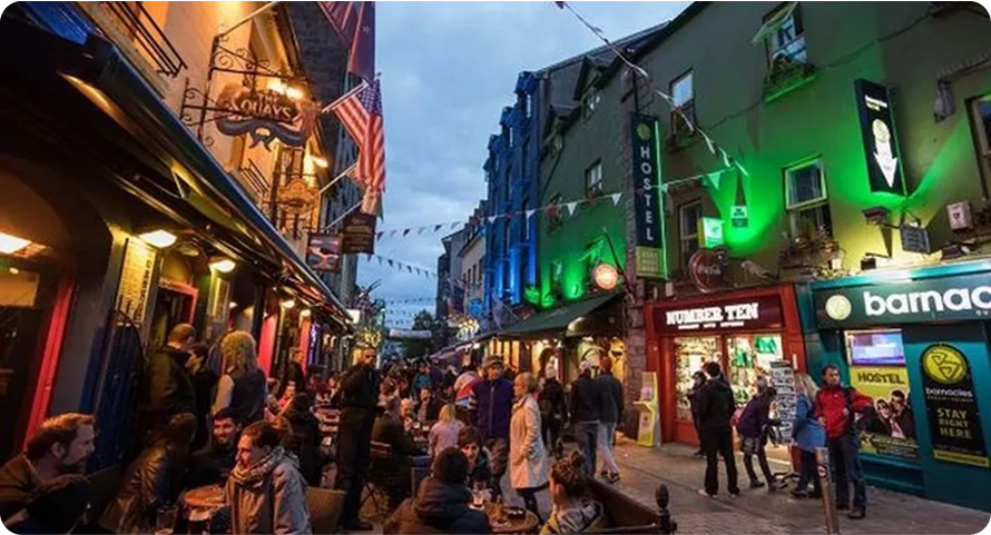 A lively street scene in the evening shows people dining and socialising at outdoor tables. Colourful buildings with bright lights and signs line the narrow street, creating a festive atmosphere.