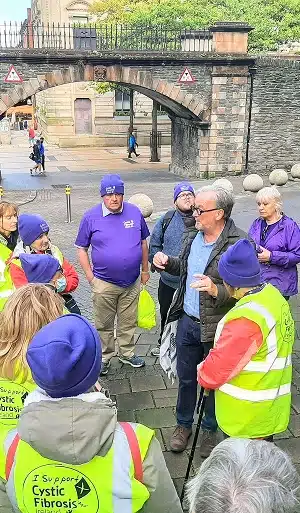 A Donegal group tours outing shows people in bright yellow vests and purple hats—some reading "I Support Cystic Fibrosis"—gathered outdoors as a man addresses them near a stone bridge and historic building.