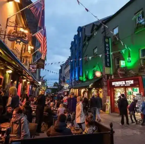 A lively street scene with people dining outdoors at cafes and bars in the evening, under fairy lights and flags, evokes the charm of a Legends of Ireland and Scotland tour amid colourful buildings and glowing signs.