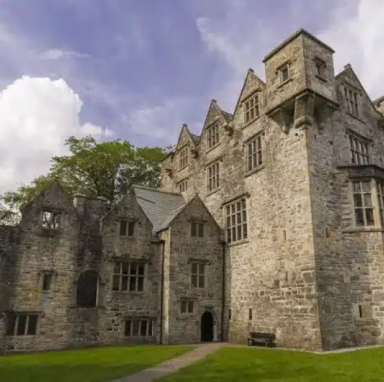 A large, historic stone castle with multiple gabled rooftops and arched windows stands beneath a partly cloudy sky—an iconic stop on the Legends of Ireland and Scotland tour—surrounded by green grass and a path leading to the entrance.