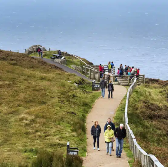 People walk along a paved path with wooden railings on a grassy cliffside overlooking the sea during the Legends of Ireland and Scotland tour. Several groups pause to admire the coastal view on an overcast day.