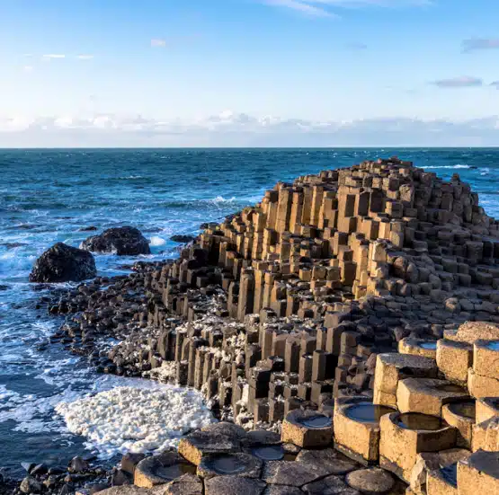 A scenic view of the Giant’s Causeway in Northern Ireland, featured on the Legends of Ireland and Scotland tour, shows natural hexagonal basalt columns beside the sea with waves crashing against the rocky shore under a blue sky.