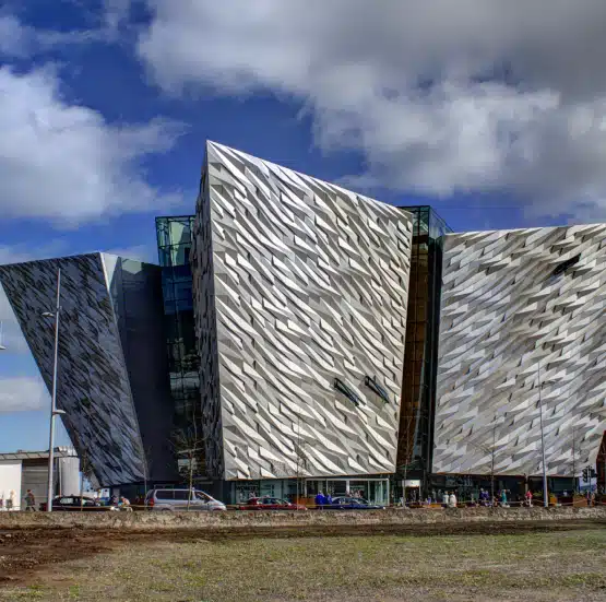 A modern building with angular, metallic façades resembling ship hulls stands beneath a partly cloudy sky. Visitors on the Legends of Ireland and Scotland tour mingle near the entrance, whilst grass covers the foreground.