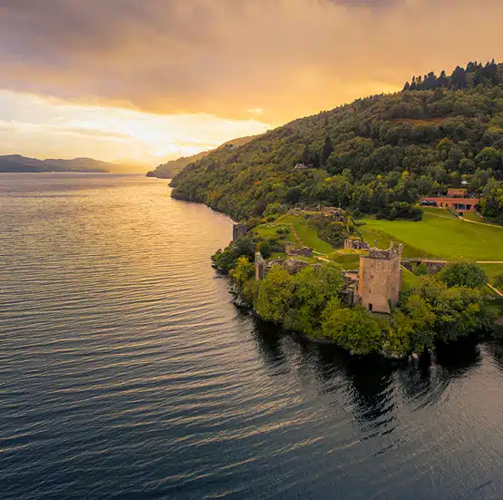 Aerial view of a ruined castle on a green hill by the edge of a large lake, with calm water and a golden sunset sky—the perfect scene from the Legends of Ireland and Scotland tour.