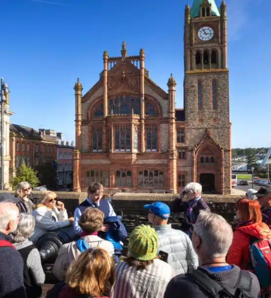 A tour guide speaks to a group of people outdoors in front of a historic red-brick building with a clock tower under a clear blue sky during the Northern Ireland highlights tour.