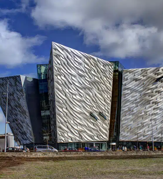 A modern building with angular, silver-coloured walls designed to resemble ship hulls, featuring textured, zigzag patterns and large glass sections under a partly cloudy sky—an iconic stop on a Northern Ireland highlights tour. Cars and people are near the entrance.