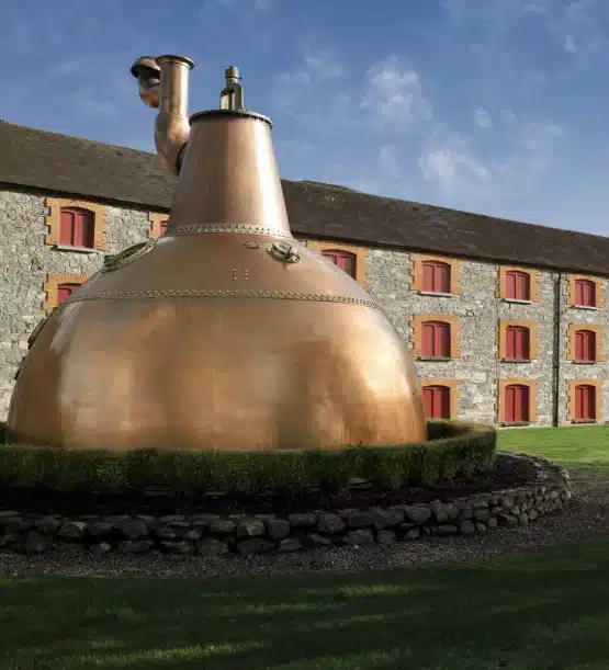 A large copper pot still sits on a circular stone base outside a stone building with red window shutters, under a clear blue sky—a classic sight on any Southern Ireland highlights tour.