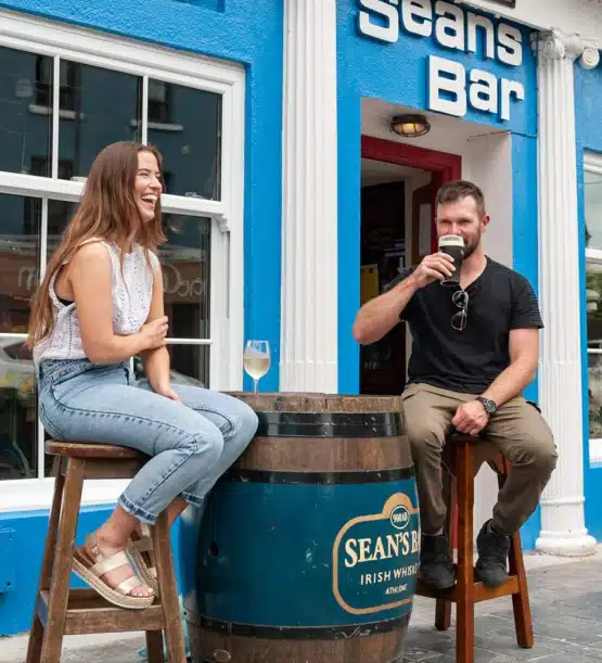 A woman and a man sit outside Sean's Bar, chatting and drinking during their Southern Ireland highlights tour. The woman smiles with her drink whilst the man, in sunglasses, enjoys a pint at a barrel table in front of the blue and white pub.