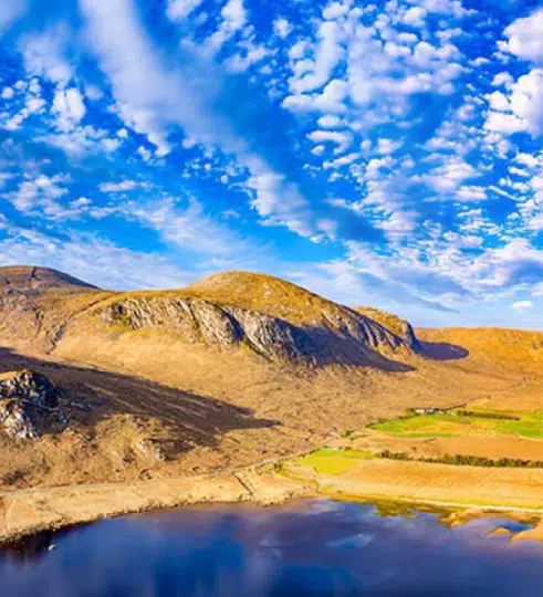 A vibrant landscape showing rugged hills under a blue sky with streaky white clouds, golden-brown fields, patches of green farmland, and a lake in the foreground—capturing the spirit of an Ireland music and dance tour.