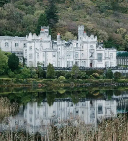 A large, white castle-like building with towers and crenellated walls stands by a calm lake, reflecting its image—an enchanting scene often admired on an Ireland music and dance tour—surrounded by lush greenery and trees on a hillside.