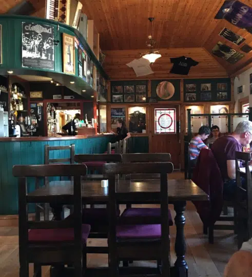 A cosy pub with wooden floors and ceiling, green bar, framed photos, music memorabilia, and several people sitting at tables—perfect for an Ireland music and dance tour. Purple cushions adorn the chairs amid vintage decor lining the walls and shelves.