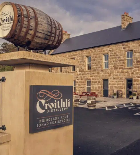A stone building with a sign reading "Croithlà Distillery" and a whisky barrel mounted above the entrance, with picnic tables and barrels outside—an inviting stop on any Ireland music and dance tour.