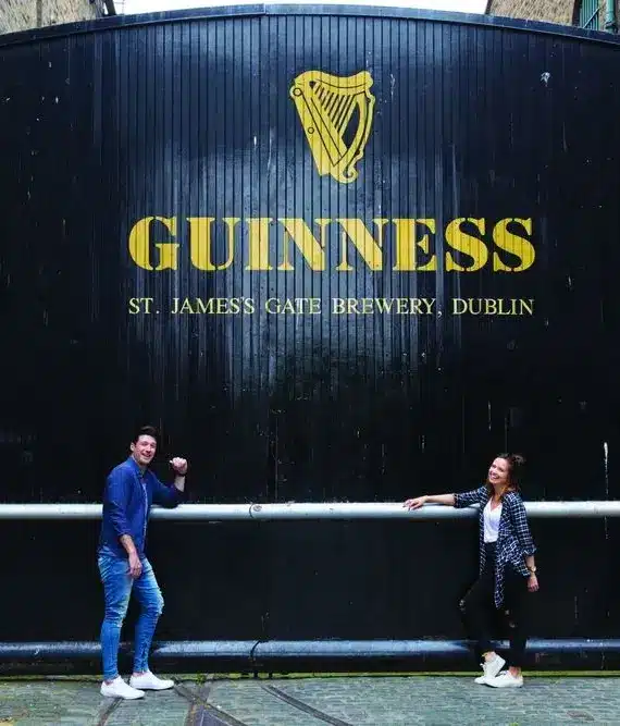 Two people pose and smile in front of a large black gate with the gold Guinness harp logo and text reading "St James's Gate Brewery, Dublin.