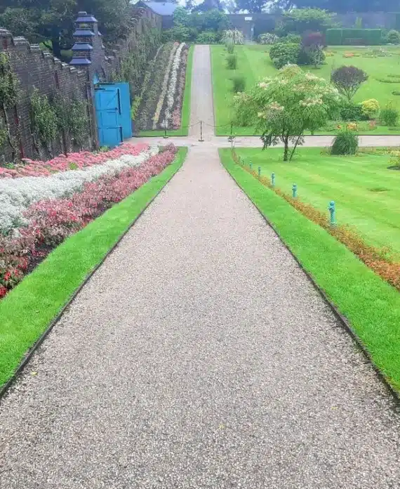 A straight gravel path bordered by neatly mown grass and colourful flower beds leads through a formal garden with trimmed bushes and a brick wall on the left.