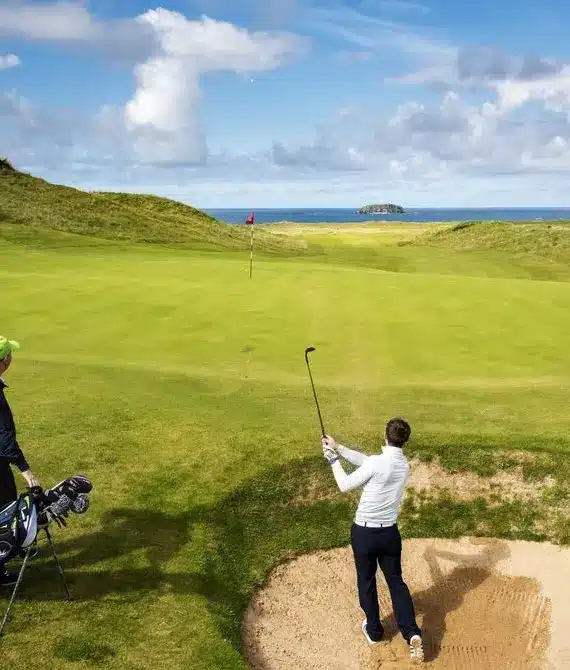 Two golfers on a green course by the sea; one stands near a golf bag while the other swings from a sand bunker towards the flag on a sunny day with clouds in the sky.