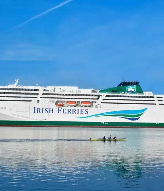 A large white ferry labelled "Irish Ferries" sails on calm blue water under a clear sky, with a small rowing boat and four people in the foreground.