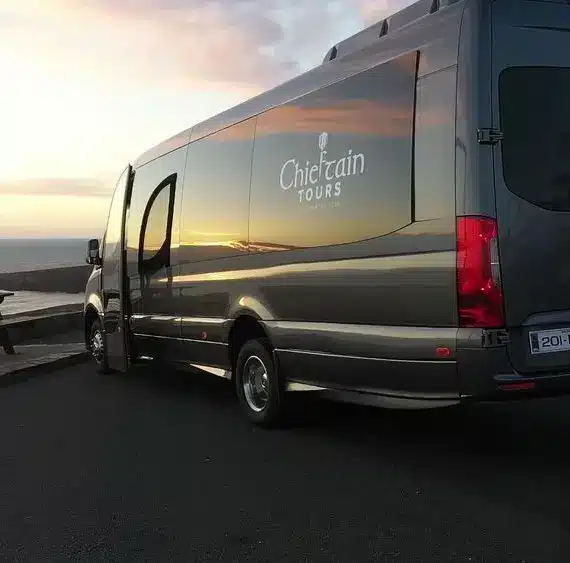 A dark grey Chieftain Tours van is parked near a wooden picnic table overlooking the sea at sunset, with soft light reflecting on the van's surface.