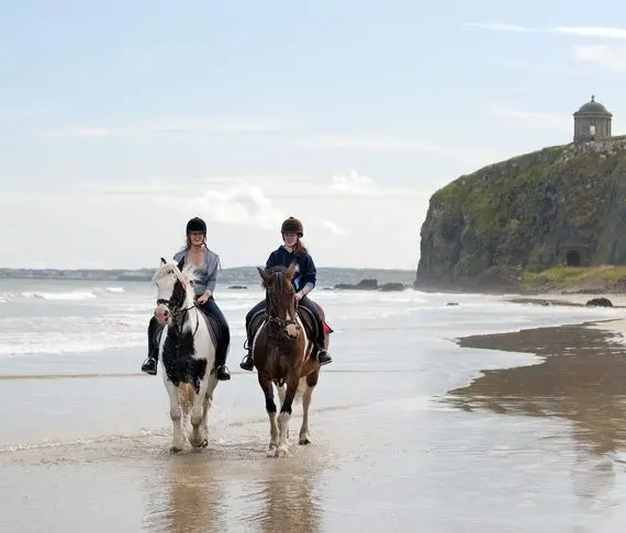 Two people ride horses along a sandy beach near the water’s edge, with cliffs and a historic stone building on a hill in the background. A few people walk and play by the sea on a partly cloudy day.