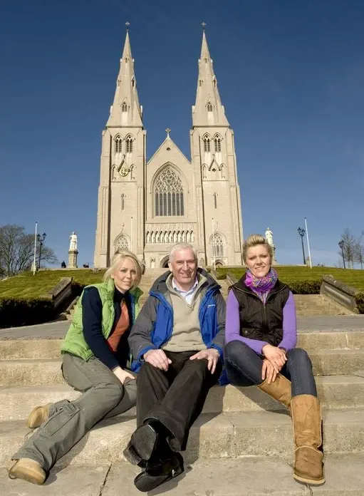 Three people sit on stone steps in front of a large church with twin spires under a clear blue sky. The group appears relaxed and smiling, with the impressive Gothic-style building behind them.
