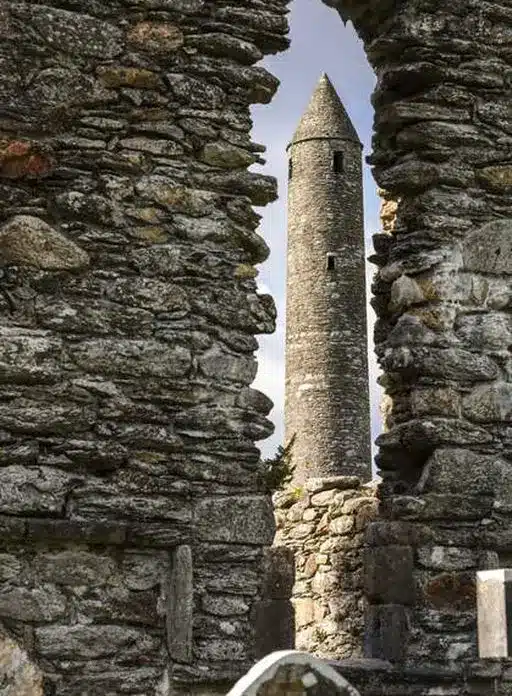 A tall, round stone tower is seen through the ruins of an old stone wall, framing the tower in the distance on a partly cloudy day.
