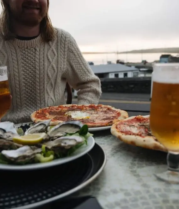 A person sits outdoors by the water, holding a glass of beer. On the table are two pizzas, a plate of oysters with lemon, another glass of beer, and a slice of brown bread.