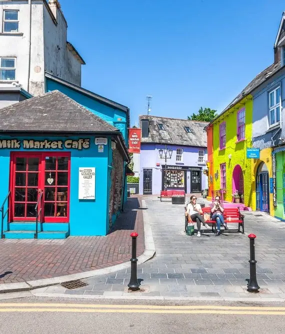 A bright, sunny street scene with colourful buildings, including a blue café called "The Milk Market Café." Several people sit on benches near vibrant yellow and green buildings, creating a cheerful atmosphere.