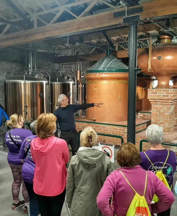 A group of people listen to a guide explaining large copper and metal stills inside a distillery, with brick and metal pipes visible in the industrial-style interior.