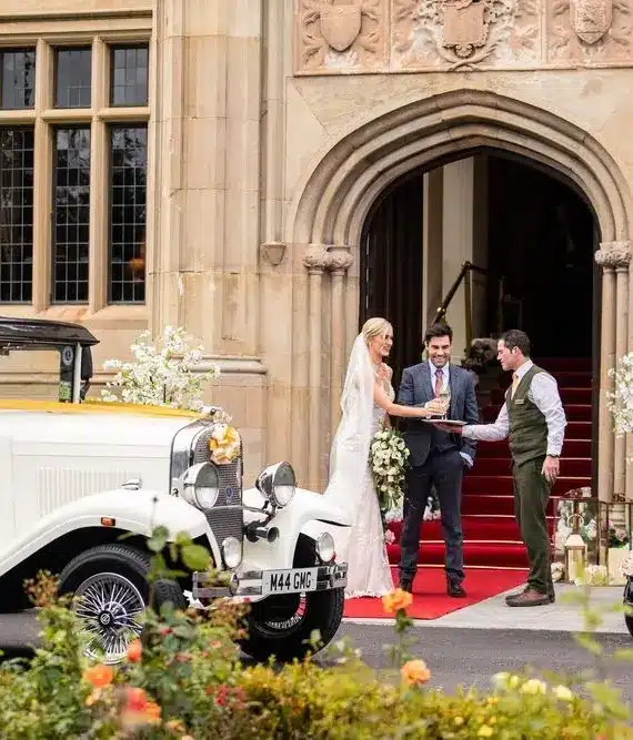 A bride and groom stand with an officiant outside a grand stone building with arched entrance, red carpet, and floral decorations, beside a vintage white car.