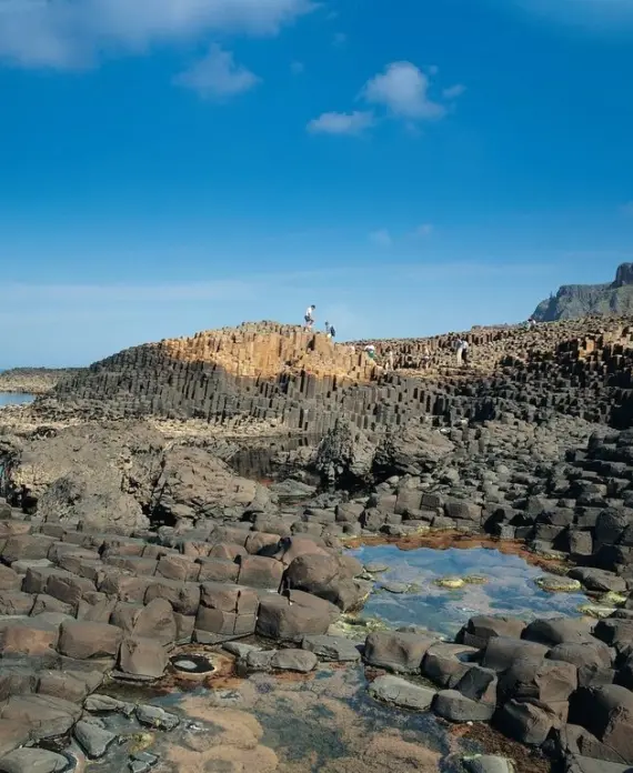 Hexagonal basalt rock columns rise from the shore at Giant’s Causeway in Northern Ireland, with a few people walking on the formations under a bright blue sky with scattered clouds.