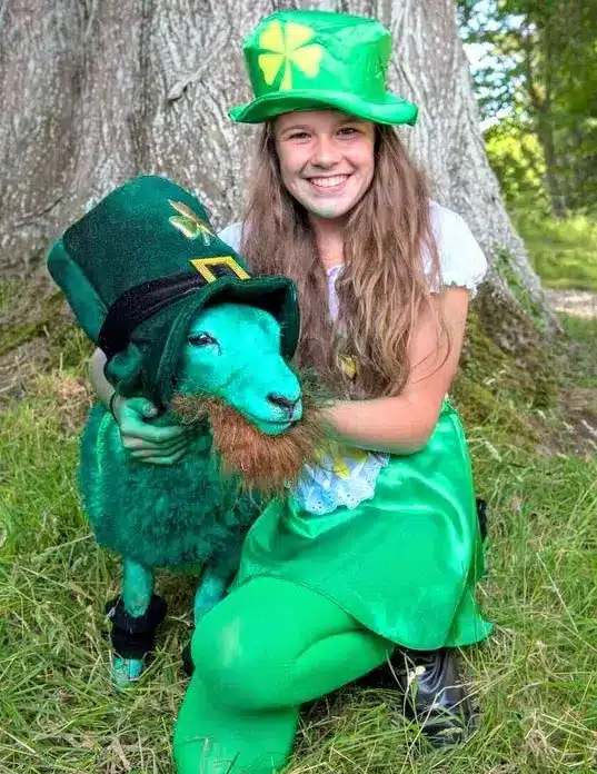 A smiling girl in green St. Patrick’s Day attire kneels beside a sheep dyed green, also dressed in a green hat and a fake orange beard, both posing cheerfully outdoors by a large tree.