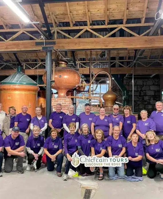 A group of people wearing purple shirts pose and smile inside a distillery with copper stills and tanks in the background. They hold a “Chieftain Tours: Discover the Edge” banner.