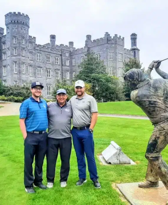 Three men in golf attire stand on grass near a golf swing statue, with a large stone castle in the background under a cloudy sky.