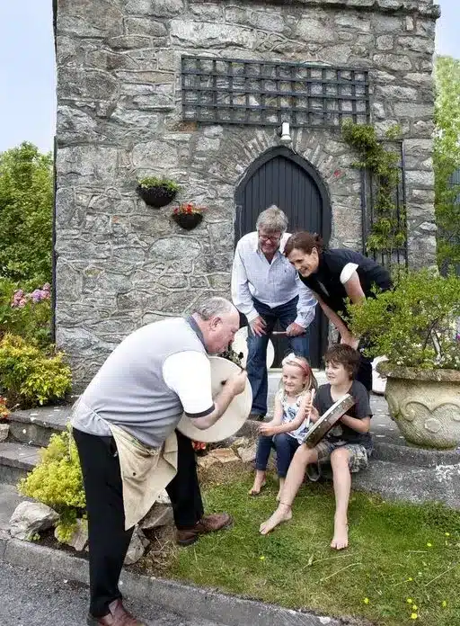 A man in an apron shows a white animal, possibly a goat or sheep, to two smiling children sitting on the grass, while two adults stand behind them, all in front of a stone building with greenery and flowers.