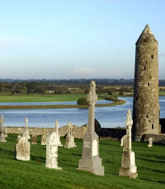 Old stone gravestones and crosses stand on a grassy hill overlooking a round mediaeval stone tower near a winding river, with fields and trees in the background under a clear sky.