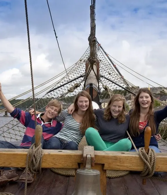Four smiling young people sit on the deck of a wooden boat, waving with one arm raised. The group appears happy, with water, buildings, and a cloudy sky in the background.