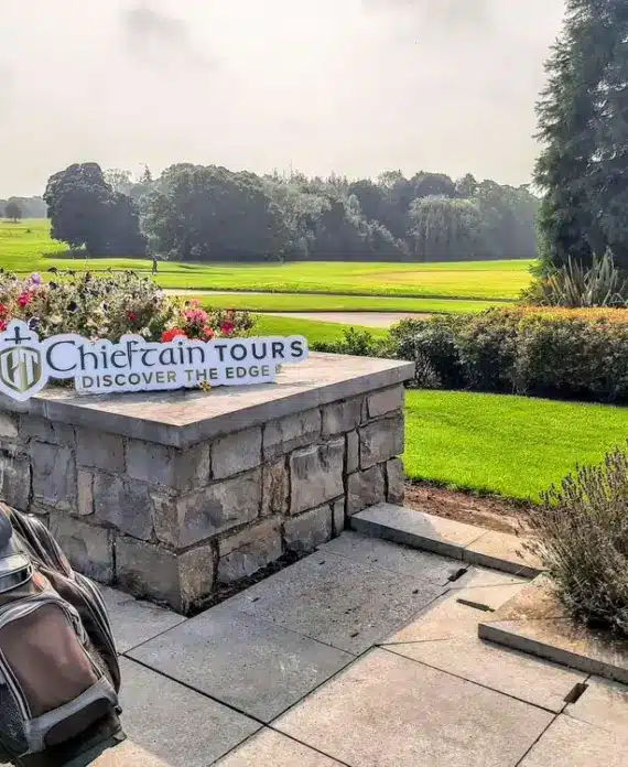 A golf bag sits on a paved patio near a stone wall with a "Chieftain Tours - Discover the Edge" sign. Behind are neatly trimmed lawns, flowerbeds, trees, and a golf course under a partly cloudy sky.