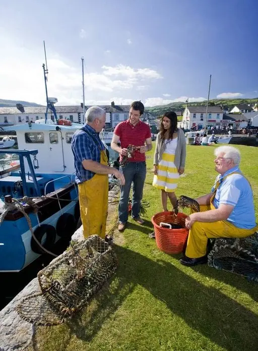 Two fishermen in yellow overalls show lobsters to a man and woman near a moored boat, with lobster pots and a red basket nearby. Buildings and a grassy area are in the background.