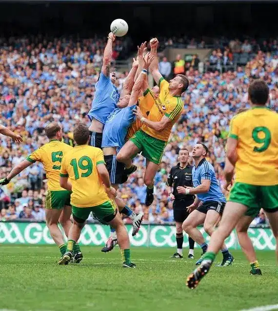 Gaelic football players in blue and yellow-green shirts jump and compete for the ball near the goal, while a crowd watches from the stands. A referee in a white jacket stands nearby.